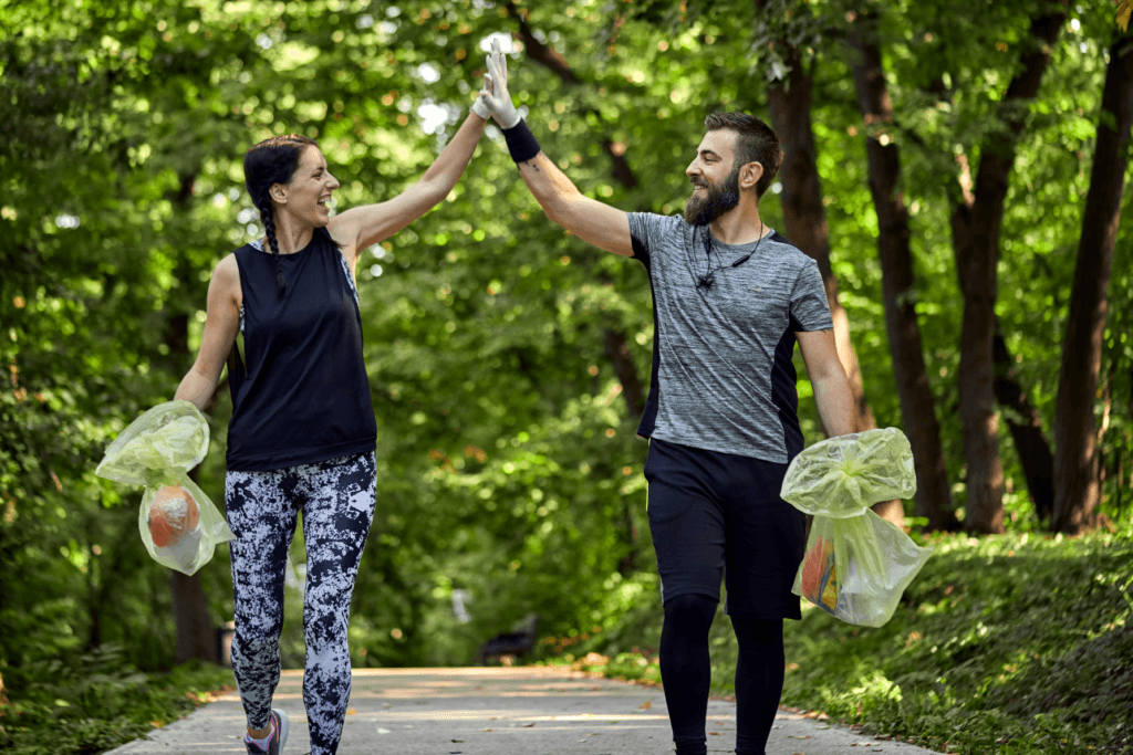 A man and woman high-fiving during a plogging activity, holding bags of collected trash, surrounded by a lush green forest.